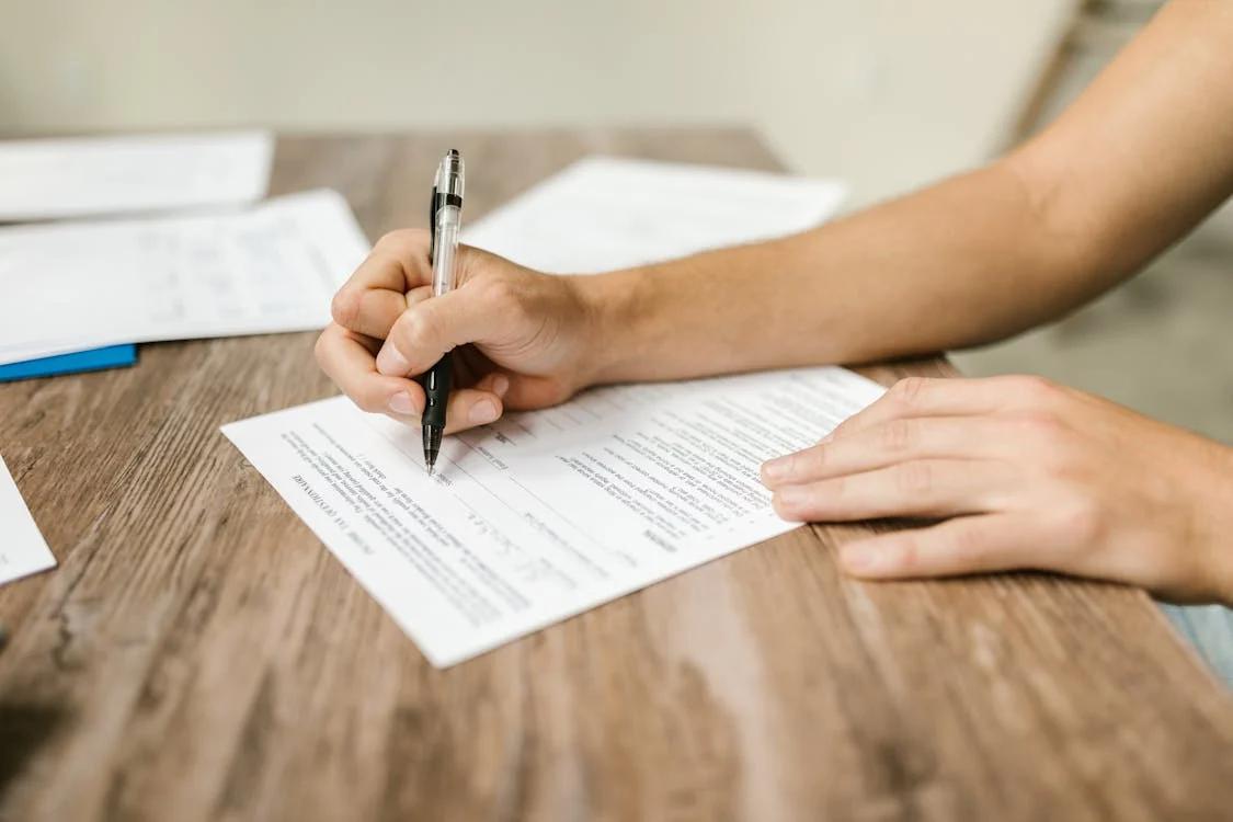 a person holding a pen, filling out forms on a wooden table