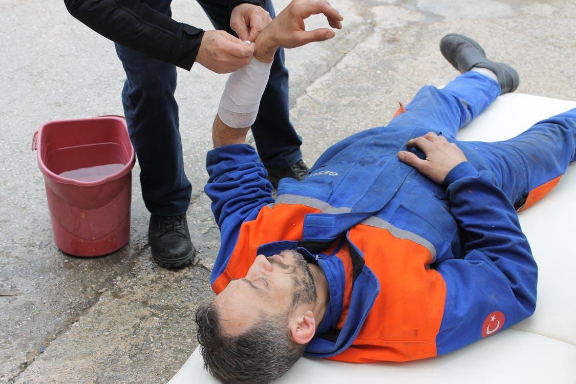 A photo showing a man in a blue and orange uniform laying on the ground and receiving bandages on their arm.
