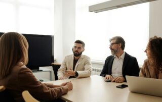 Four people are seated on a desk with their laptops open.