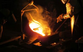 Metal manufacturing workers operating industrial equipment while wearing protective gear in a fabrication facility.